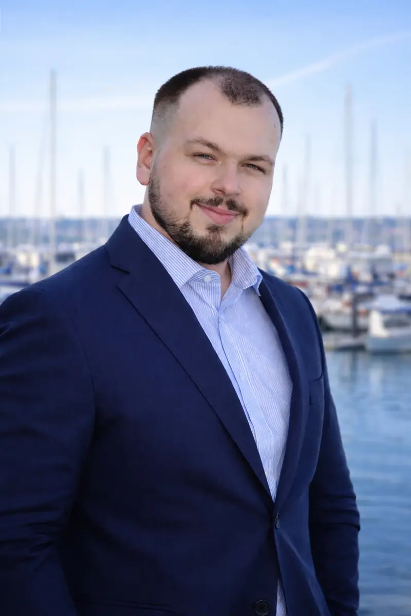 Max Petrov in a navy blazer, smiling, with sailboats behind him
